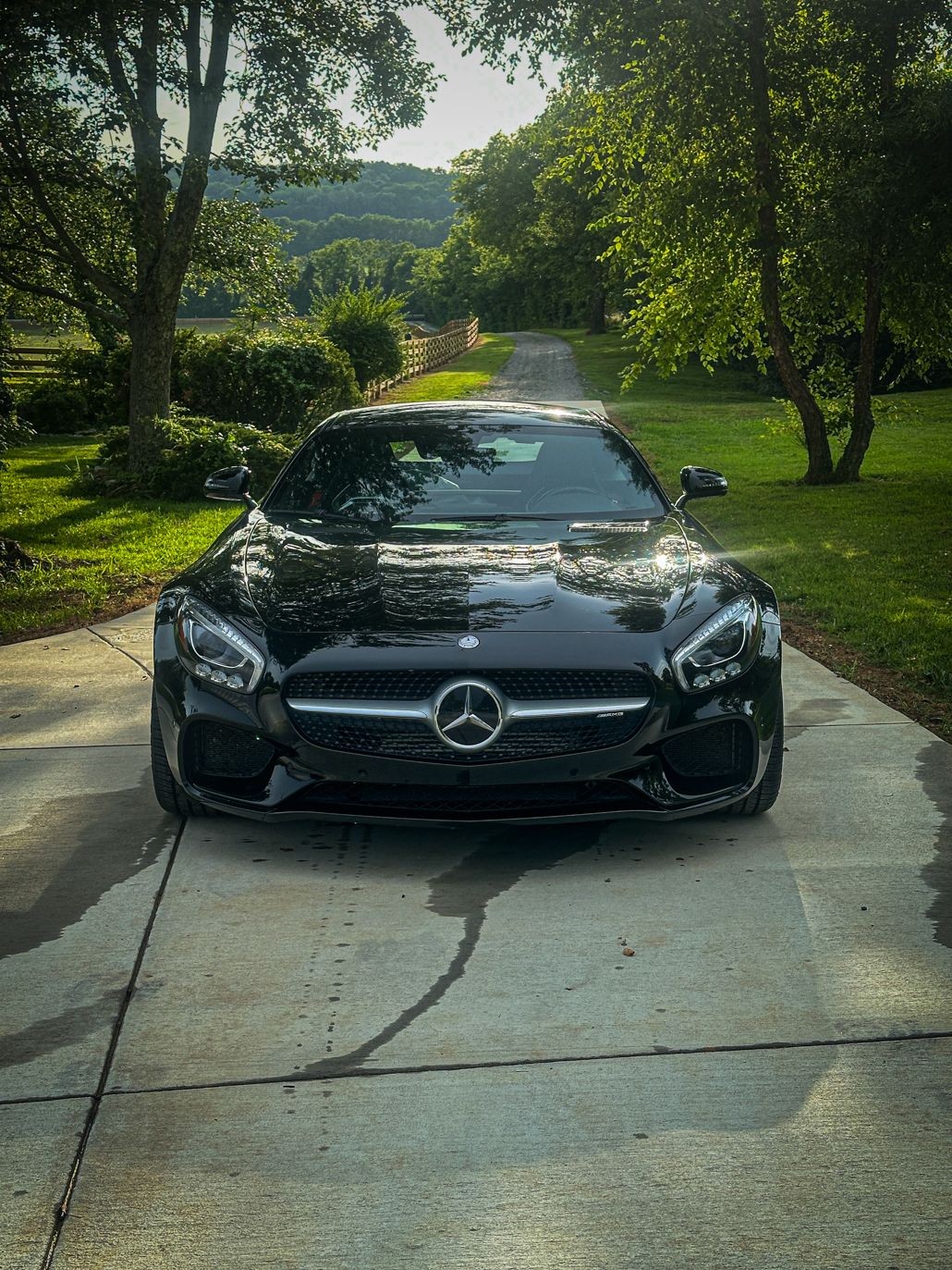 Black luxury car parked on a driveway surrounded by green trees and a scenic hilly background.